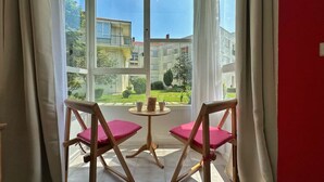 Een kamer met twee houten stoelen met roze kussens, een klein rond tafeltje en uitzicht op gebouwen en groen buiten via een groot raam.