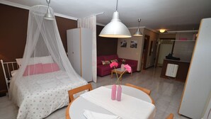 A bedroom with a bed, a bedside table, a round table with chairs, a wardrobe, a sofa, and a kitchenette area.