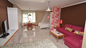 A living room with a pink sofa, a TV, a dining table with chairs, a coffee table, and a bed with a canopy.