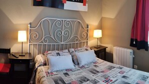 A neatly made bed with a newspaper print bedspread, two bedside lamps, a decorative headboard, and a red curtain. - Estudio en la Playa del Sable - El Corrillo 5602 (Arnuero)