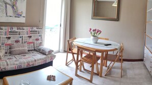 A cozy room with a sofa covered in newspapers, a round dining table with chairs, a potted plant, and a mirror on the wall. - Estudio en la Playa del Sable - El Corrillo 5602 (Arnuero)