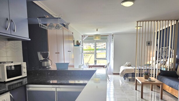 A modern kitchen with a central island, a dining area with a sofa, and a view through a glass door. - Loft en Isla Playa - El Corrillo 5604 (Arnuero)