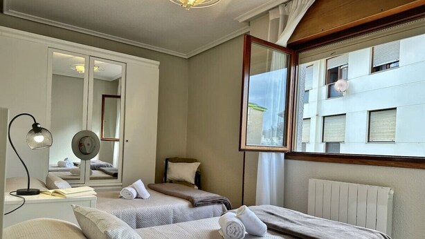 Una habitación de hotel con dos camas, un espejo, una mesita de noche con lámpara y un ventanal con vista a edificios.