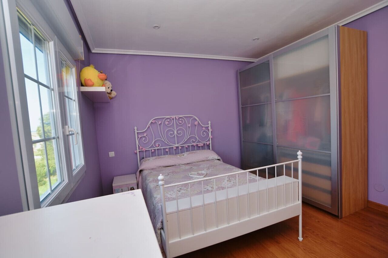 Une chambre à coucher avec un lit en métal blanc, des murs violets, une grande fenêtre et une porte-fenêtre coulissante.