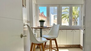 Una cocina con armarios blancos, una ventana con vistas a palmeras y dos sillas blancas.