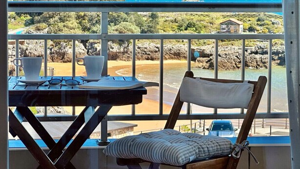 Un balcón con mesa y silla, con vista a una playa y un litoral rocoso.
