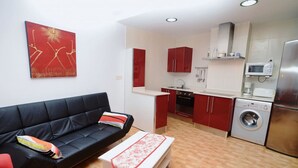 A compact living space with a black sofa, a red accent wall, a kitchen area with red cabinets, a washing machine, and a microwave. - Bajo con vistas inigualables - La Caseta 5907 (Isla)