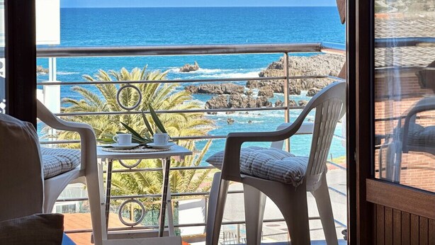 A balcony with two white chairs, a small table, and a view of the ocean and rocky coastline. - Apartamento en playa de Ris - Pineda 6607 (Noja)
