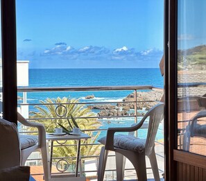 Un balcon avec deux chaises blanches, une petite table et une vue sur l’océan et la côte rocheuse.