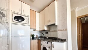 A compact kitchen with a washing machine, a microwave, and wooden cabinets. - Apartamento en playa de Ris - Pineda 6607 (Noja)