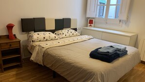 Ein ordentlich bezogenes Bett mit einer beigen Bettdecke, ein Nachttisch mit einer roten Vase, ein an der Wand montierter Fernseher und ein Fenster mit Jalousien.