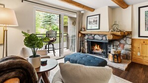 A cozy living room with a stone fireplace, a large window, and a balcony with outdoor furniture. - 700 Monarch Condos by iTrip Aspen Snowmass (Aspen)