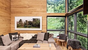 Un intérieur moderne avec un plafond en bois, un canapé agrémenté d’un plaid blanc moelleux, une table basse et une grande fenêtre donnant sur un paysage boisé.