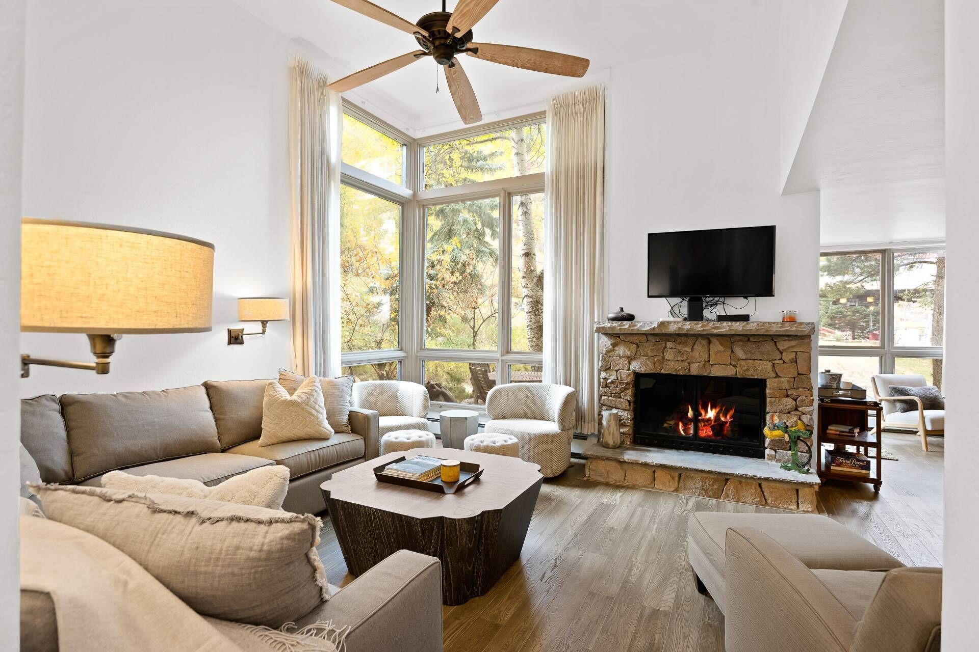 A spacious living room with a stone fireplace, large windows, a ceiling fan, and a flat-screen TV mounted above the fireplace.