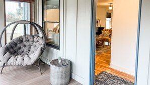 A room with a grey upholstered armchair, a wicker side table with a plant, and a view into an adjacent room with a patterned rug and a black chair. - Welcome Race Fans! - Oversize Parking - Firepit - Mural - West Indy - Brownsburg (Indianapolis)