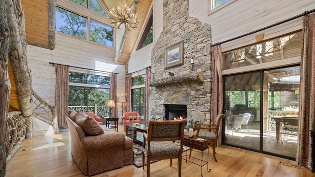 A spacious living room with a stone fireplace, wooden flooring, and large windows offering a view of the outdoors.