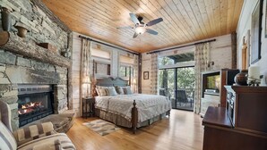 A cozy bedroom with a stone fireplace, a large bed, a ceiling fan, and a television.