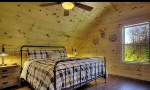 Une chambre dans une cabane en bois, avec un lit en métal, une table de chevet, un ventilateur de plafond et une fenêtre donnant sur la forêt.
