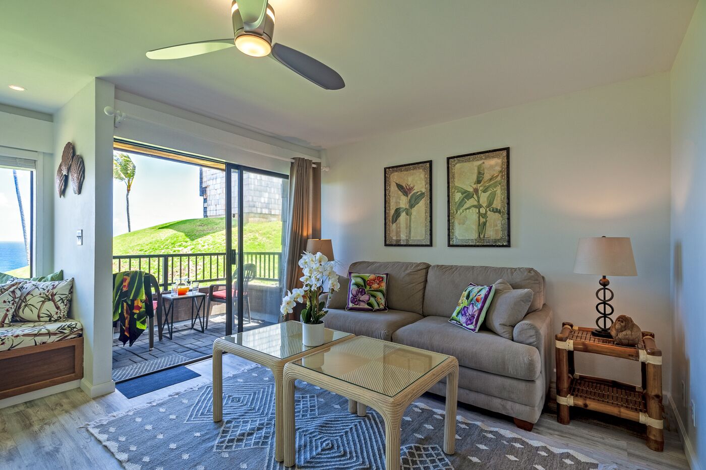 A spacious living room with a glass door leading to a balcony, a sofa set, a coffee table, and a small side table with a lamp.
