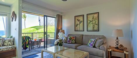 A spacious living room with a glass door leading to a balcony, a sofa set, a coffee table, and a small side table with a lamp.