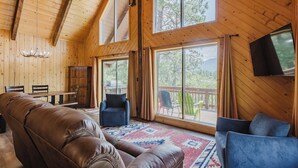 A cozy cabin interior with a leather couch, blue chairs, a wooden dining table, and a large window offering a view of the outdoors. - Close to Wolf Creek, Fish San Juan River, Escape the Crowds (Pagosa Springs)