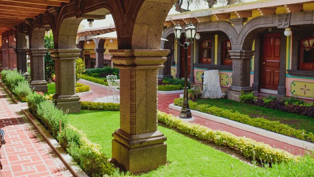 A covered walkway with stone columns and a garden area with a pathway and benches. - Gran Misiones Tequisquiapan (Tequisquiapan)