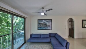 Ein modernes Wohnzimmer mit einem großen Ecksofa, einem Deckenventilator, einem gerahmten Bild an der Wand und Blick durch große Fenster.