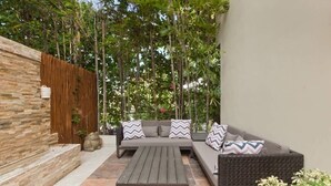 Eine Terrasse im Freien mit Steinmauer, Holzzaun, einer grauen Sofagarnitur mit gemusterten Kissen und einem Holztisch.
