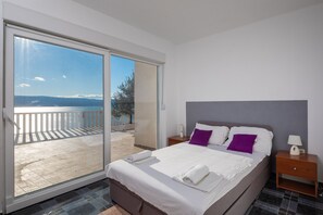 Relax in a spacious room with a large bed, a nightstand, and a sliding door leading to a balcony with a sea and mountain view.