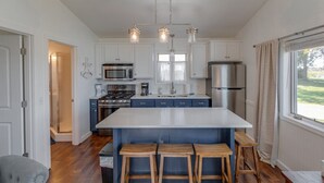 Una cocina moderna con gabinetes blancos, una isla grande con taburetes, electrodomésticos de acero inoxidable y vistas al exterior a través de una ventana.