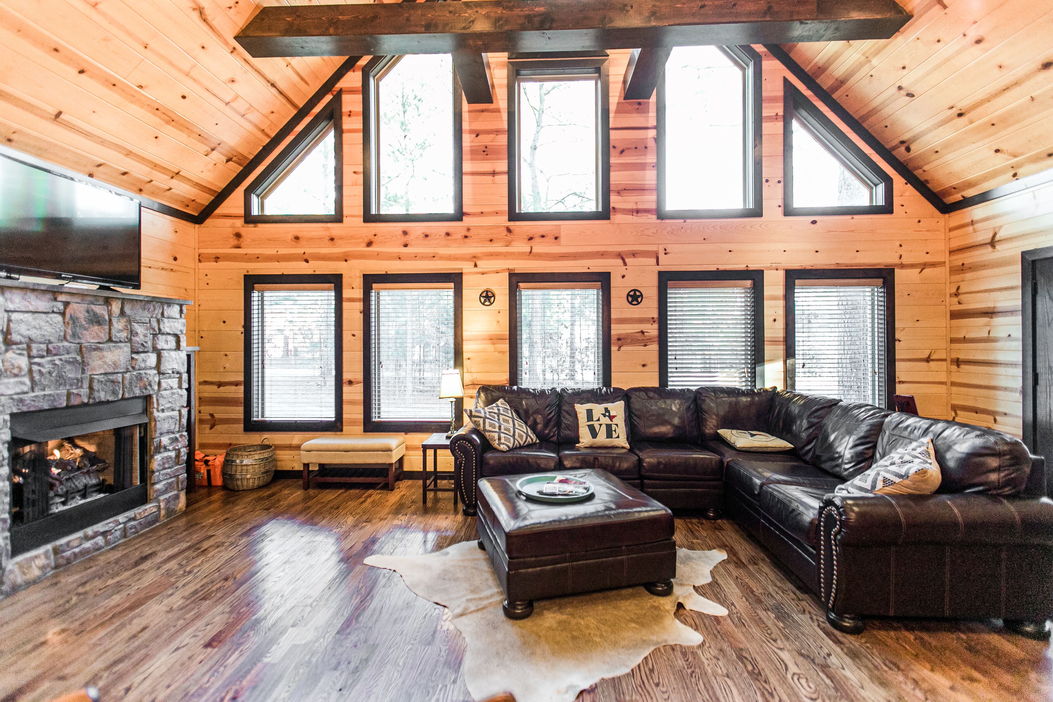 A log cabin interior with a stone fireplace, leather sofas, and large windows.