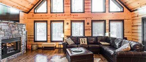 A log cabin interior with a stone fireplace, leather sofas, and large windows.