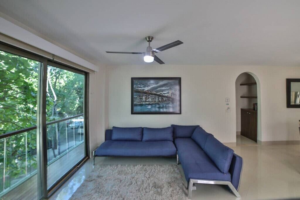 Ein modernes Wohnzimmer mit einem großen Ecksofa, einem Deckenventilator, einem gerahmten Bild und Blick durch große Fenster.