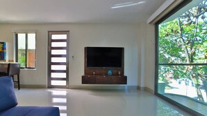 Een moderne woonkamer met een flatscreen televisie op een houten standaard, een blauwe bank en een groot raam met uitzicht op bomen.