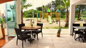 Un patio con una mesa y sillas, una puerta de cristal que da a vista de la playa, y un parque infantil al fondo.