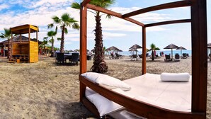Una cama de madera con almohadas blancas y estructura de madera, sobre una playa arenosa con palmeras y sombrillas de playa al fondo.