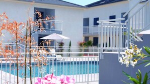Zona junto a la piscina con cielo azul despejado, piscina y edificio blanco con balcones.