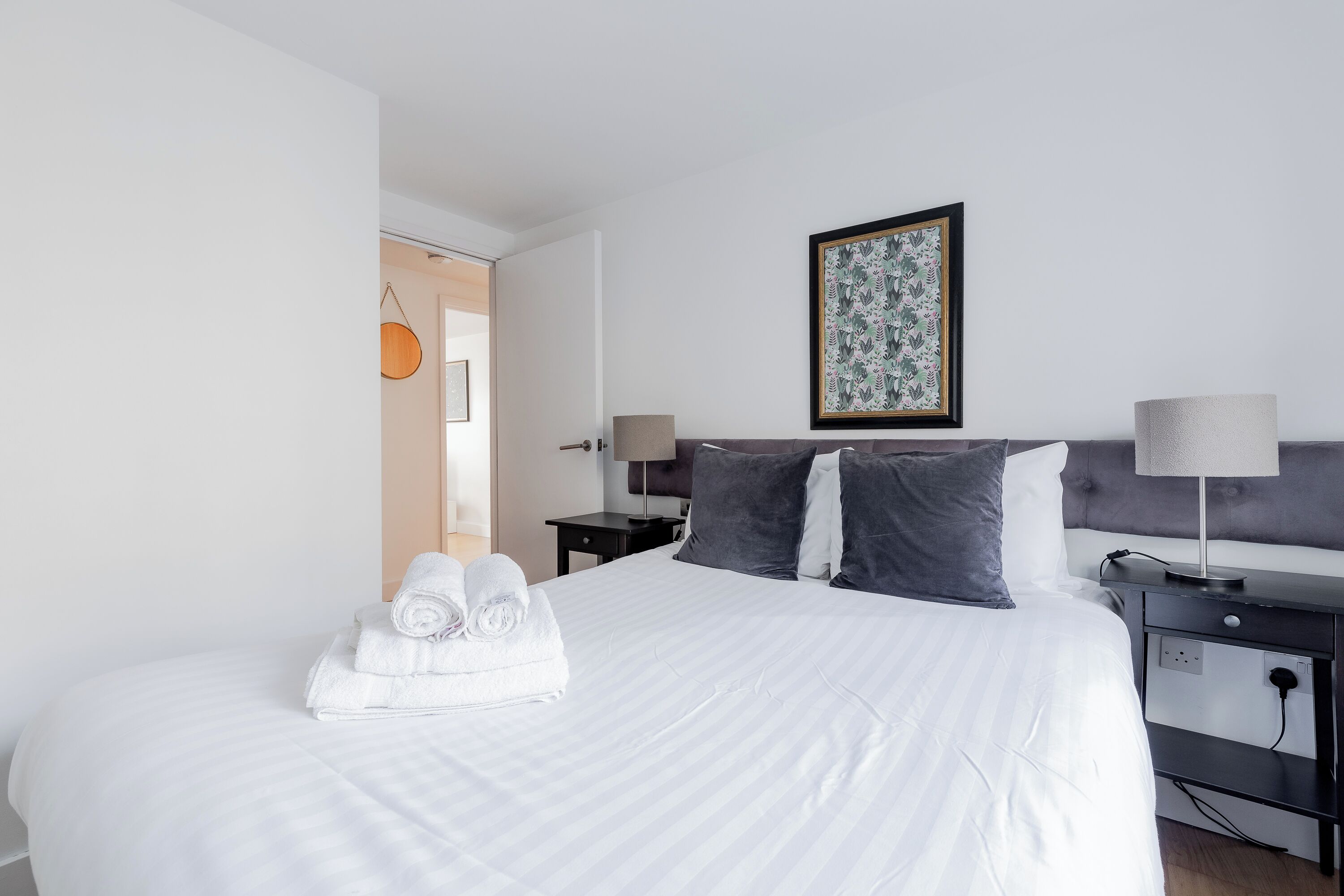 A neatly made bed with white linens and rolled towels, a grey upholstered headboard, a bedside table with a lamp, and a framed artwork on the wall.