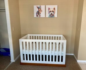 A white crib with a patterned mattress in a room with beige walls and two framed dog paintings on the wall.