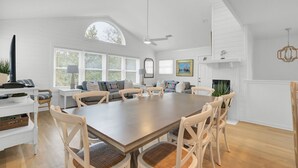 A spacious dining area with a large wooden table and white chairs, surrounded by windows with blinds, a ceiling fan, and a fireplace. - Award-Winning Resort Amenities | Pelican Bay 38 (Isle of Palms)