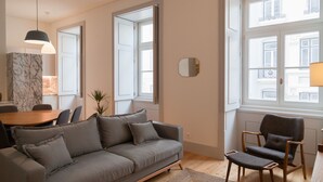 A modern living room with a grey sofa, a wooden dining table, and a window with white shutters.