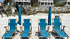 Cuatro sillas de madera en la playa con cojines azules y sombrillas a juego, sobre arena. Se observan edificios y una escalera en el fondo.