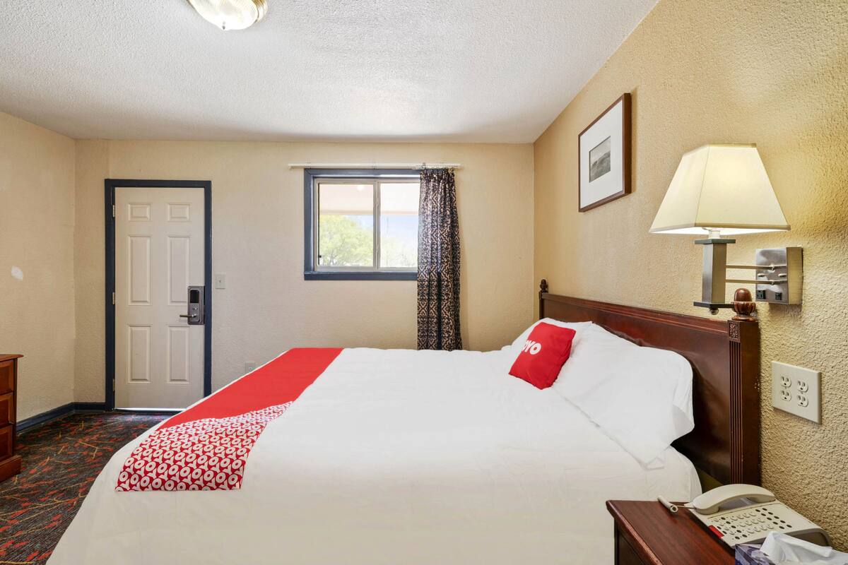A hotel room with a bed, a bedside lamp, a telephone, a window with curtains, and a door.