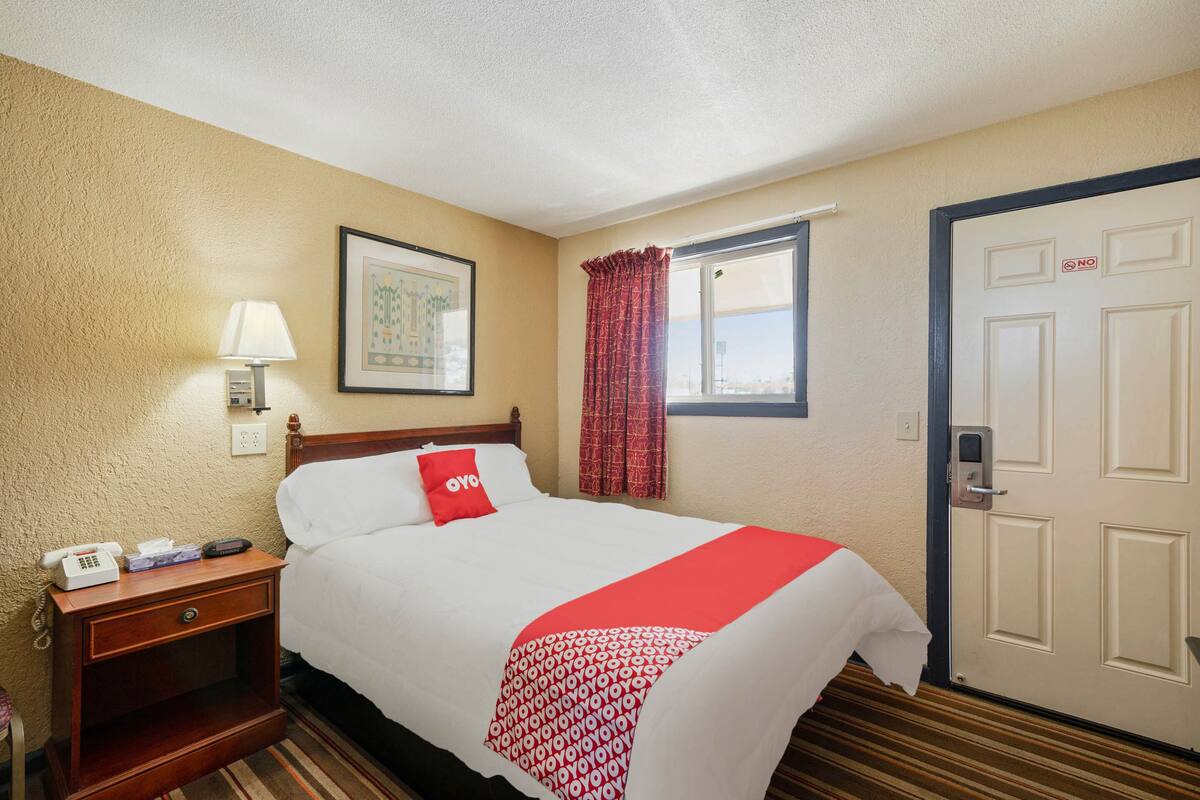 A hotel room with a bed, a nightstand with a phone and tissues, a window with red curtains, and a door with a 'NO' sign.