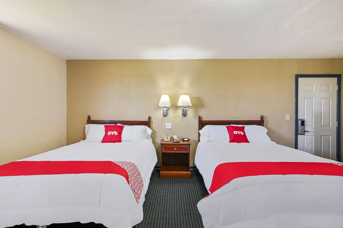 A hotel room with two beds, each with a red and white bedspread, a wooden headboard, and a nightstand with a lamp between the beds.