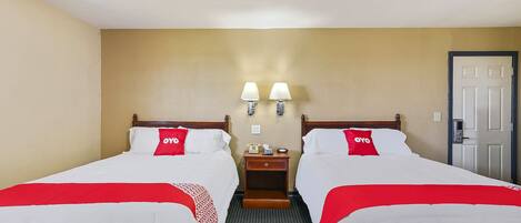 Habitación de hotel con dos camas, cada una con cubrecama rojo y blanco, cabecero de madera y mesita de noche con lámpara entre las camas.