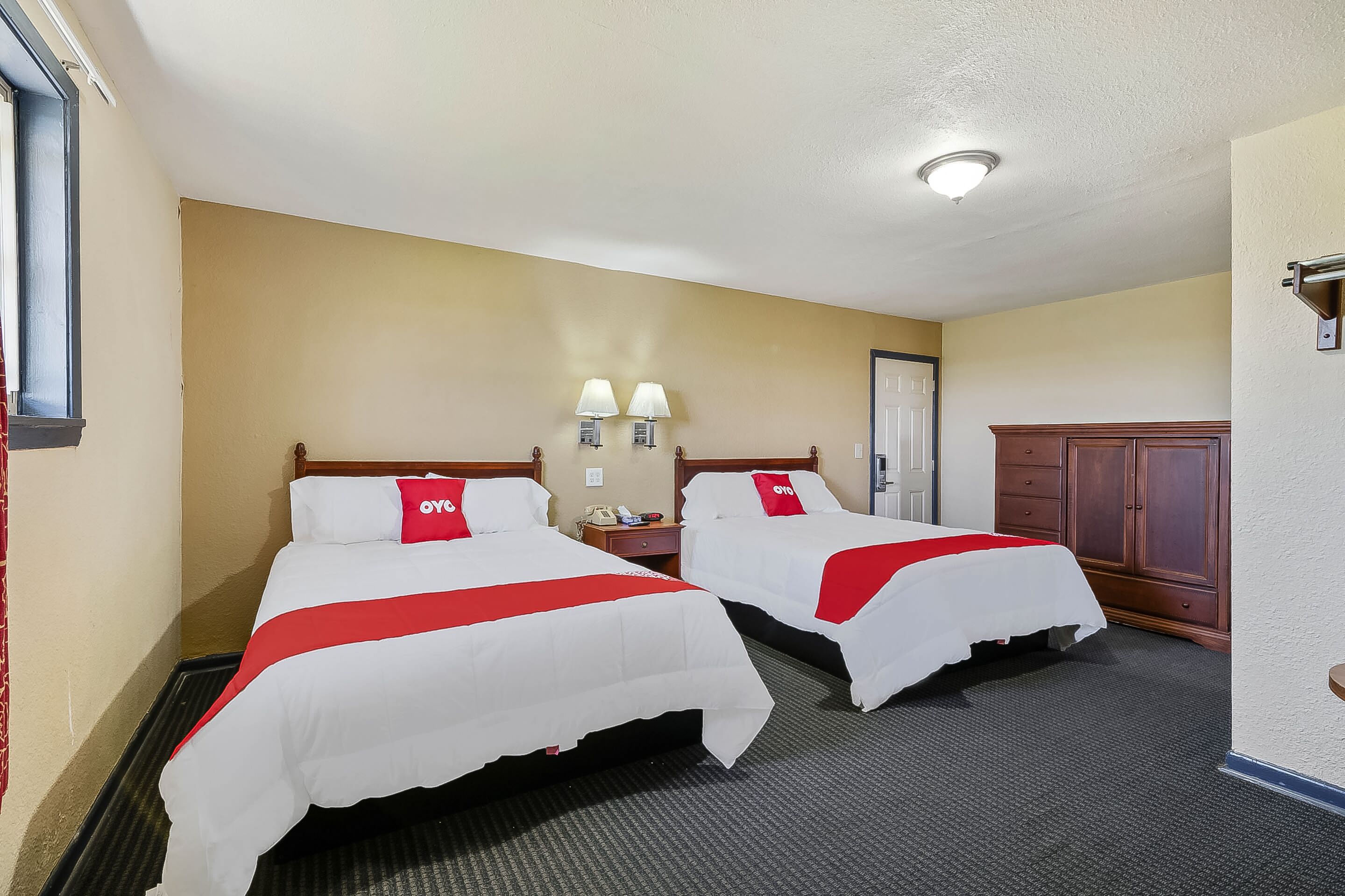 Habitación de hotel con dos camas, cada una con una colcha roja y blanca y un cojín a juego. Hay dos mesitas de noche con lámparas, un armario de madera y una puerta al fondo.