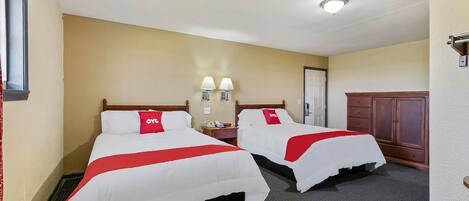 A hotel room with two beds, each with a red and white bedspread and a matching pillow. There are two bedside tables with lamps, a wooden wardrobe, and a door in the background.