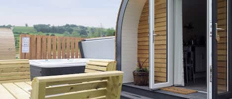 Una terraza de madera con un jacuzzi, bancos de madera y una puerta que da a una zona de cocina.