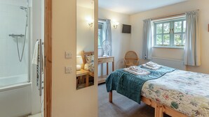 Un dormitorio con cama, una silla de madera, una mesita de noche, un espejo, una ventana con cortinas y un rincón de ducha.